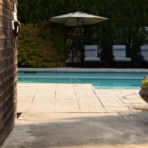 A swimming pool with three lounge chairs and a large umbrella, surrounded by greenery and a potted plant, viewed from a stone patio.