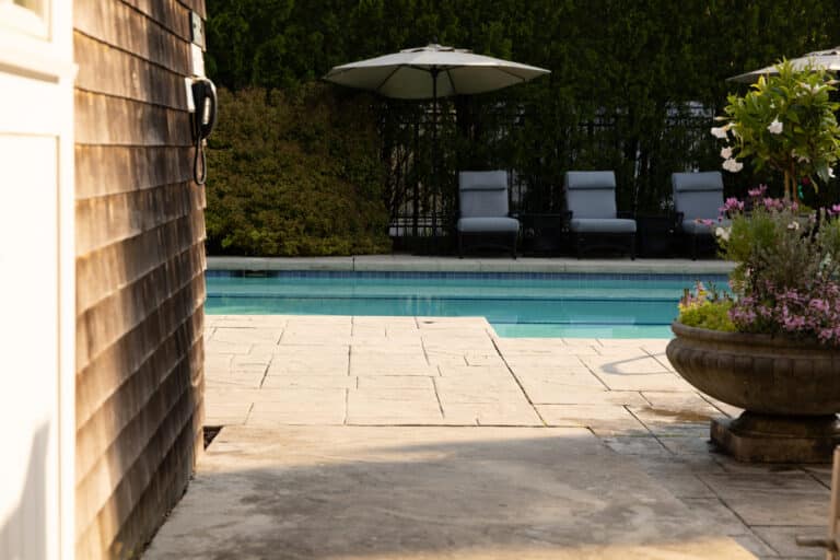 A swimming pool with three lounge chairs and a large umbrella, surrounded by greenery and a potted plant, viewed from a stone patio.