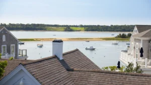 Overlapping rooftops in the foreground overlook a calm waterway with anchored boats; trees and grassy land are visible across the water under a clear sky.
