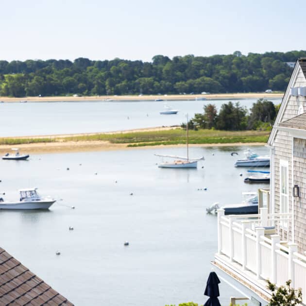 Several boats are anchored in a calm bay near a shoreline with green trees, with part of a shingle-style house and its balcony visible in the foreground.