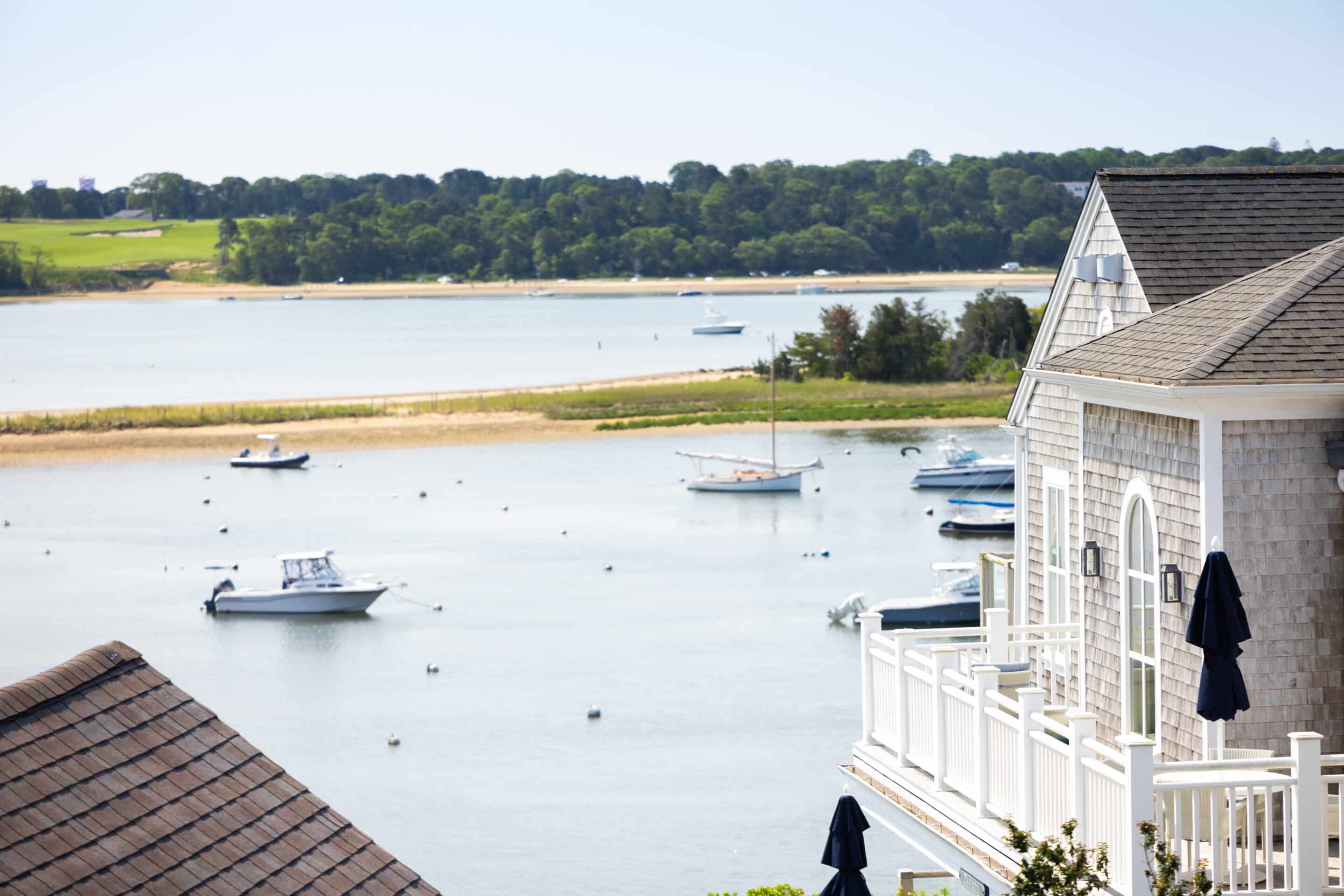 Several boats are anchored in a calm bay near a shoreline with green trees, with part of a shingle-style house and its balcony visible in the foreground.