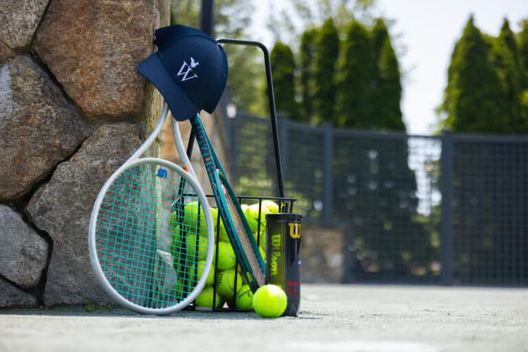 A tennis racket, basket of tennis balls, can of tennis balls, and a navy cap rest against a stone wall on an outdoor Racquet Sports court.