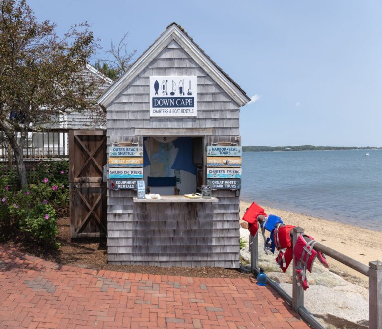 Small wooden shack labeled "Down Cape Charters & Boat Rentals" by a brick path, with life jackets hanging outside, next to a beach and water.