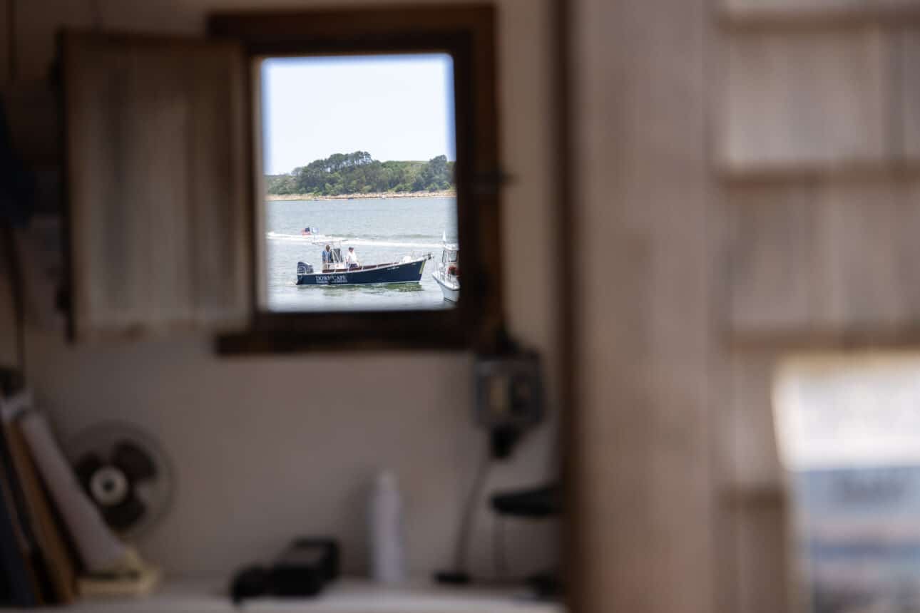 A boat with people on board is seen through a window from inside a room at Wequassett Resort; the blurred interior contrasts with the sharp focus on the boat and water outside.