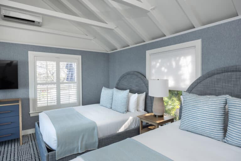 A cozy bedroom at this Cape Cod resort features two double beds with light blue bedding, a window with white shutters, a dresser with a TV, and a lamp on the nightstand between the beds.