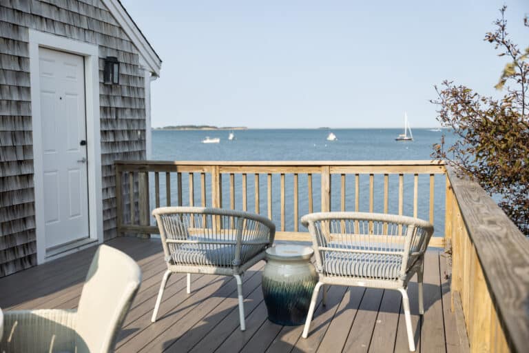 Two wicker chairs and a small table on a wooden deck at Wequassett Resort and Golf Club overlook calm water with sailboats in the distance under a clear sky.
