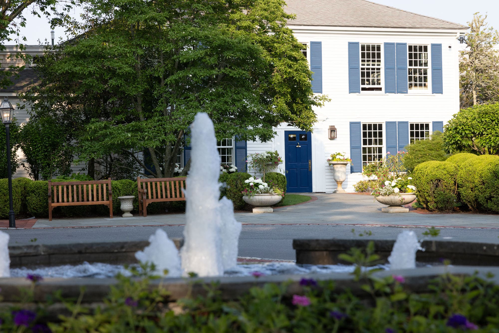 A white building with blue shutters and a blue door is seen behind a fountain, benches, and landscaped greenery on a sunny day—an inviting spot often featured on local Garden Tours.