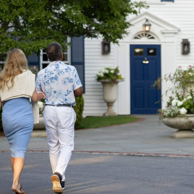 A man and woman walk arm in arm toward a white house with blue shutters, surrounded by greenery and planters.
