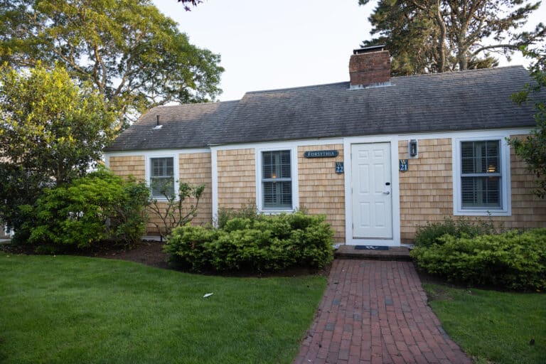 Charming single-story cottage with wood shingles, white trim, and a brick path leading to a white front door—ideal for those seeking a classic Cape Cod wedding venue surrounded by green shrubs and grass.