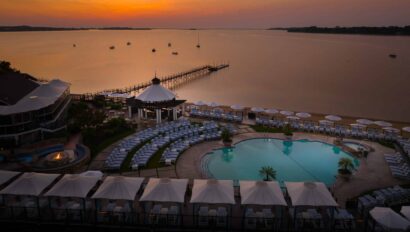 A resort pool area with lounge chairs, cabanas, and inviting poolside retreats overlooks a calm lake at sunset, featuring a pier extending into the water and boats in the distance.