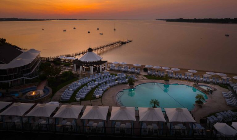 A resort pool area with lounge chairs, cabanas, and inviting poolside retreats overlooks a calm lake at sunset, featuring a pier extending into the water and boats in the distance.