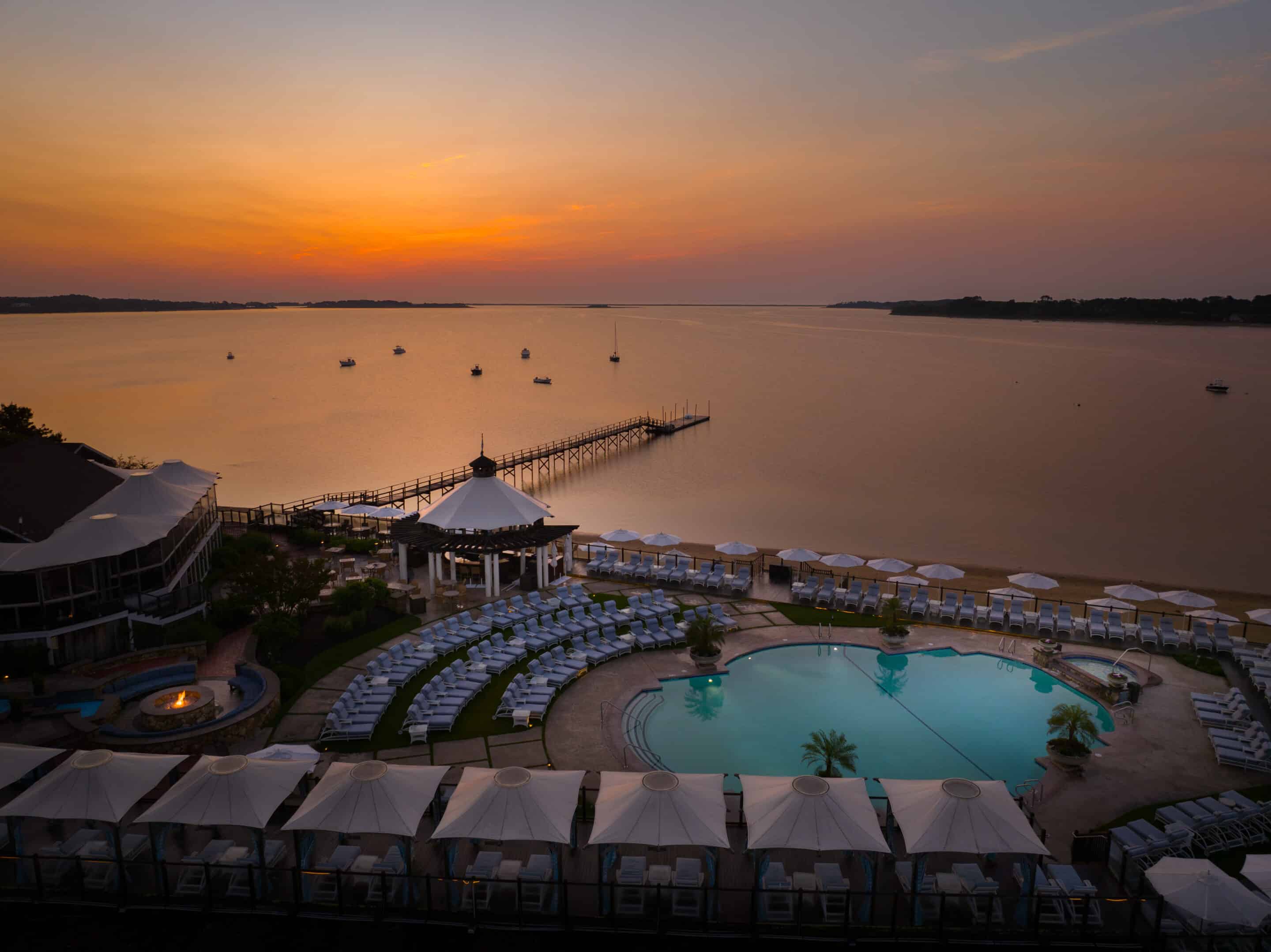 A resort pool area with lounge chairs, cabanas, and inviting poolside retreats overlooks a calm lake at sunset, featuring a pier extending into the water and boats in the distance.