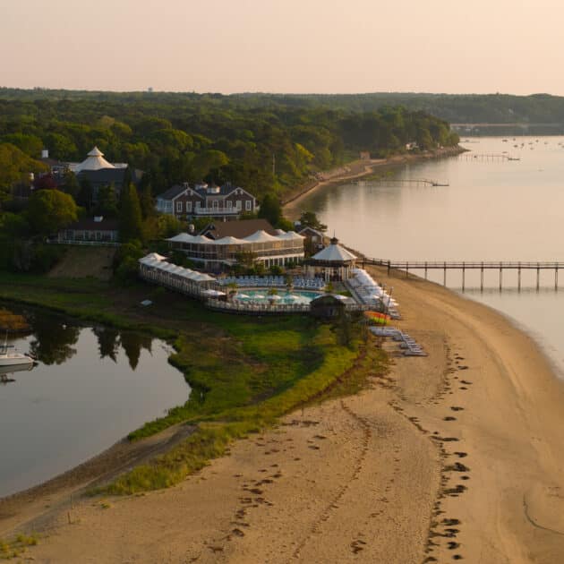Aerial view of a beachfront resort with a pool, white buildings, a sandy beach, a dock extending into calm water, and forested area in the background, taken at sunset.