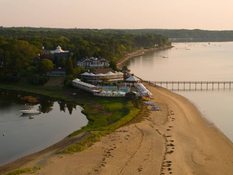 Aerial view of a beachfront resort with a pool, white buildings, a sandy beach, a dock extending into calm water, and forested area in the background, taken at sunset.