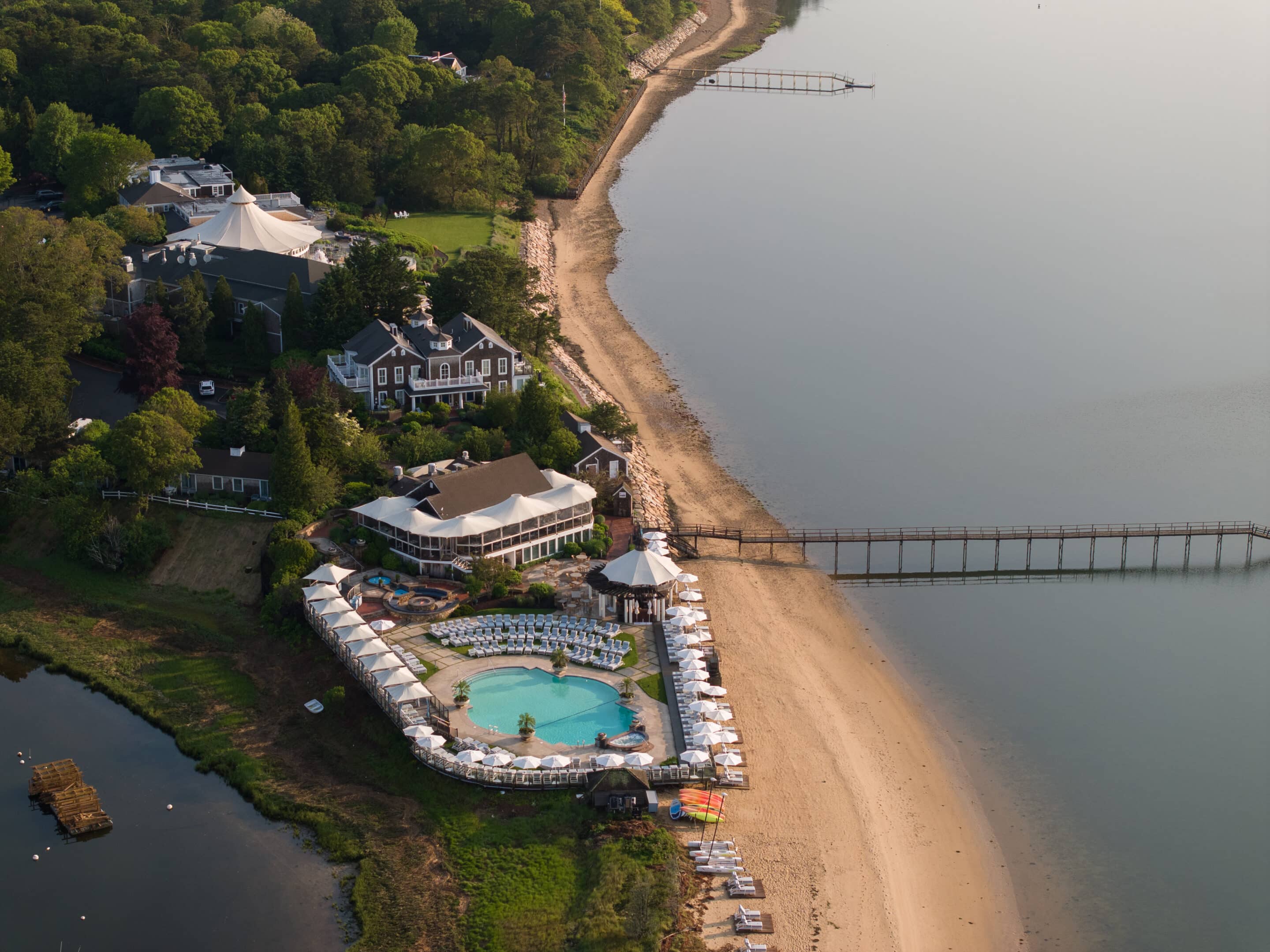 Aerial view of a sustainable resort with a pool, beach umbrellas, and a pier extending over calm water, surrounded by trees and eco-friendly buildings.