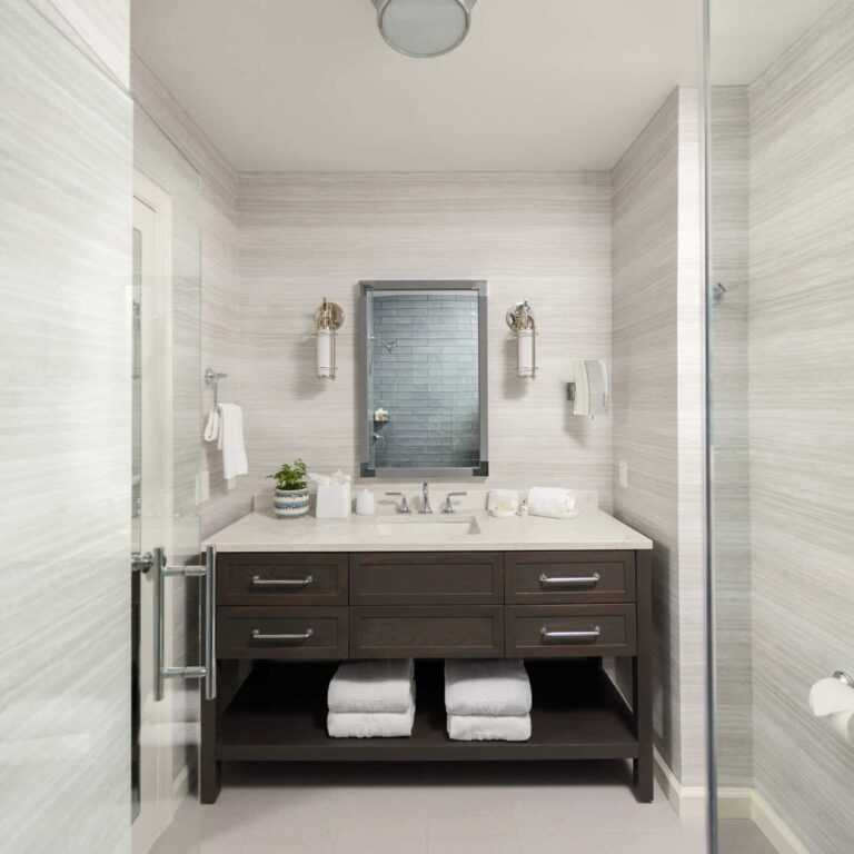 Modern bathroom at Wequassett Resort and Golf Club with a double-sink vanity, two wall sconces, a mirror, rolled towels under the sink, and a glass door on the left.