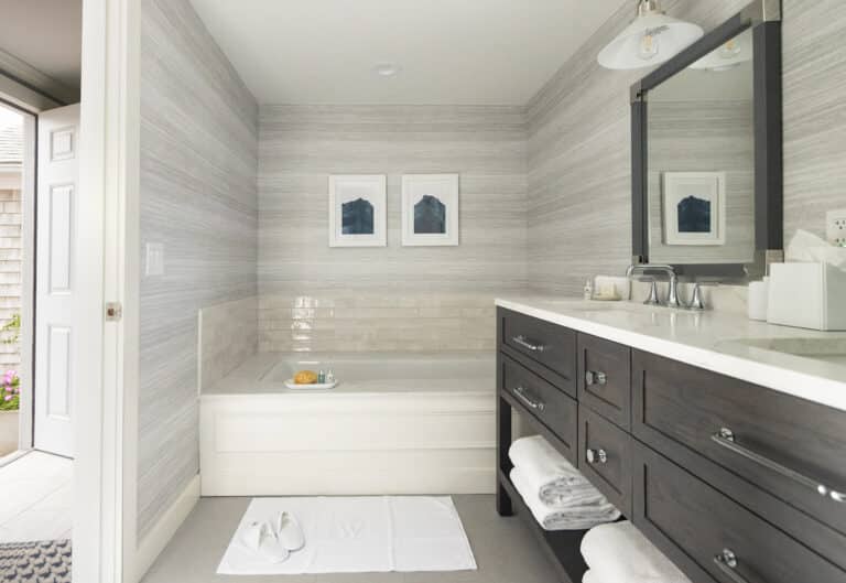 Modern bathroom at a Cape Cod resort with a built-in bathtub, two framed prints on the wall, dark wood vanity with double sinks, and white towels neatly stacked on shelves. Door to outside open.