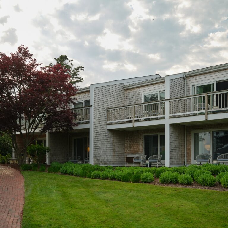 Two-story building with wood siding and balconies, surrounded by green lawn, shrubs, and a red brick pathway under a cloudy sky—an inviting Cape Cod hotel near 28 Atlantic and premier Cape Cod golf courses.