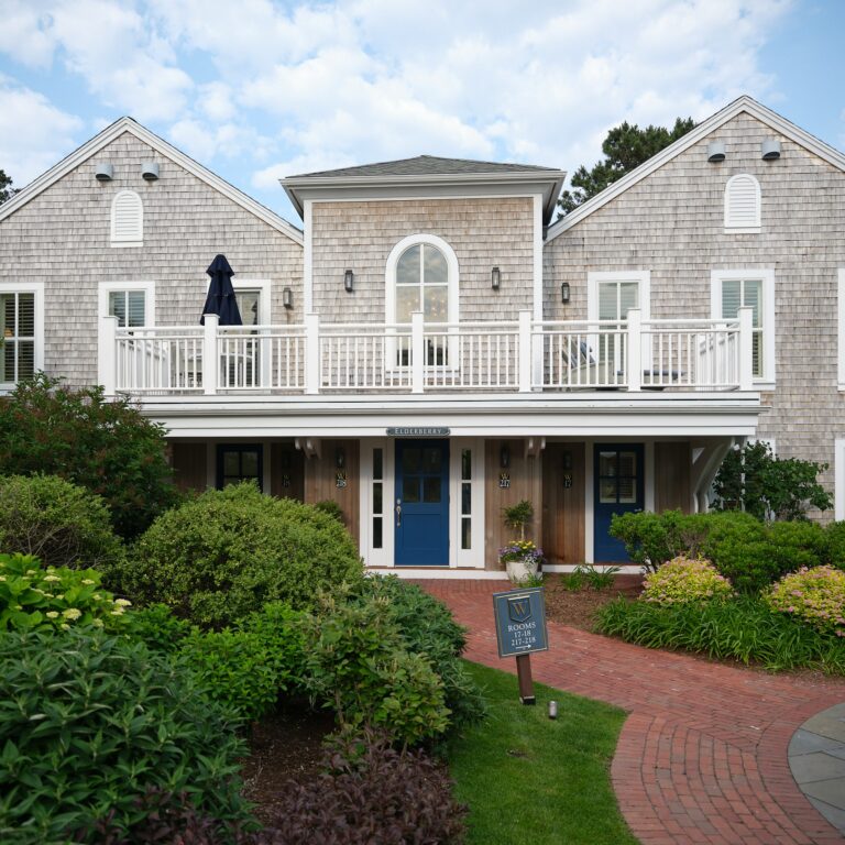 A two-story gray shingle building with white trim and blue double doors, this charming Cape Cod resort is surrounded by landscaped greenery and a brick walkway leading to the entrance.