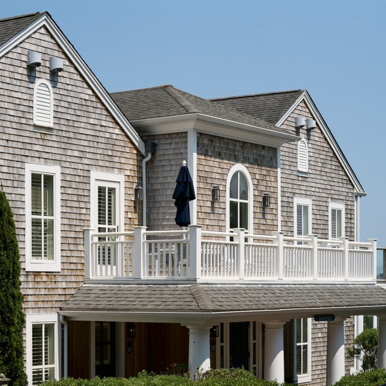 A two-story house with gray shingle siding, white trim, large windows, and a second-floor balcony featuring a closed navy umbrella—perfect as an intimate Cape Cod wedding venue or retreat near your favorite Cape Cod golf course.