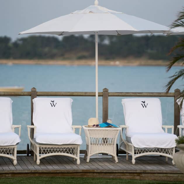 Four white lounge chairs with towels and umbrellas face a stunning waterfront view at Wequassett Resort, with a small table, hat, and book between them. A potted plant is visible on the right.
