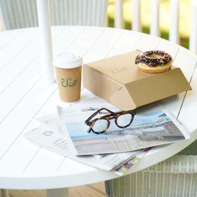 A white round table with a cardboard coffee cup, a closed takeout box, a chocolate donut with sprinkles, newspapers, and a pair of glasses on it.