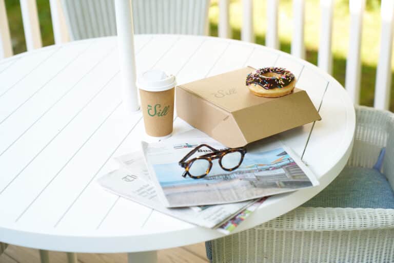 A white round table with a cardboard coffee cup, a closed takeout box, a chocolate donut with sprinkles, newspapers, and a pair of glasses on it.