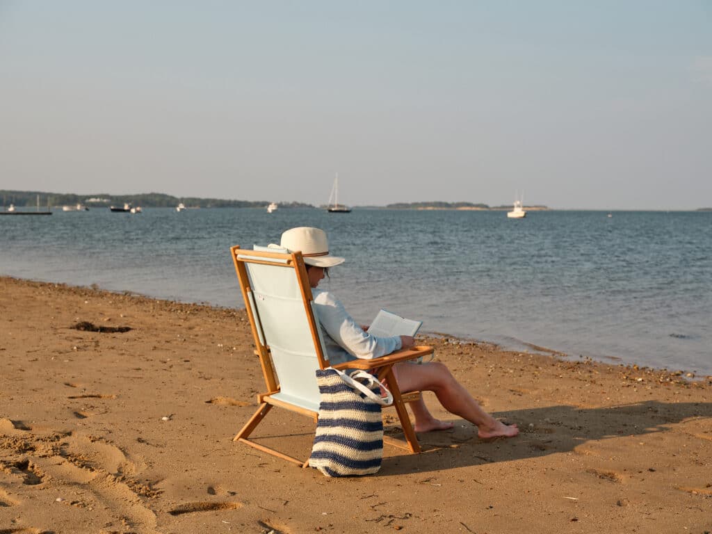 Person sitting in a beach chair on sandy shore, reading a book with a striped bag nearby; sailboats and calm water in the background.