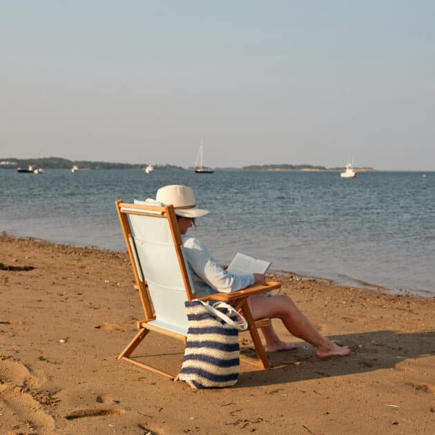 Person sitting in a beach chair on sandy shore, reading a book with a striped bag nearby; sailboats and calm water in the background.