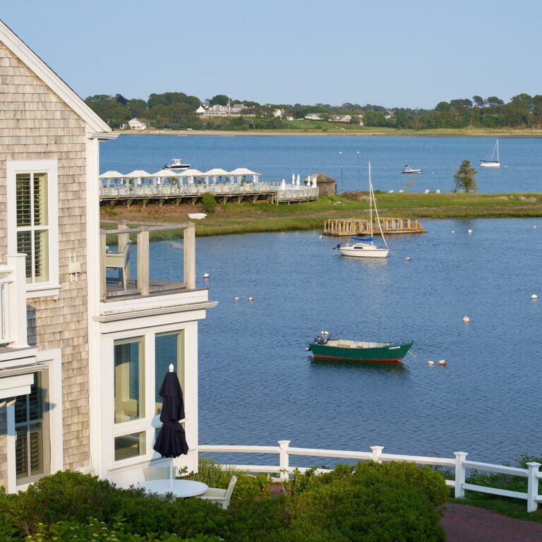 A waterfront house overlooks a calm bay dotted with sailboats, much like the serene views near 28 Atlantic, a renowned Cape Cod wedding venue, with distant buildings glimmering across the water under a clear sky.