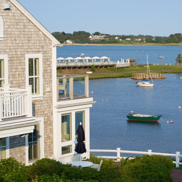 A wooden shingle house overlooks a calm body of water near a dock, with boats and white cabanas opposite—a serene scene reminiscent of a Cape Cod hotel or the peaceful vistas by 28 Atlantic.
