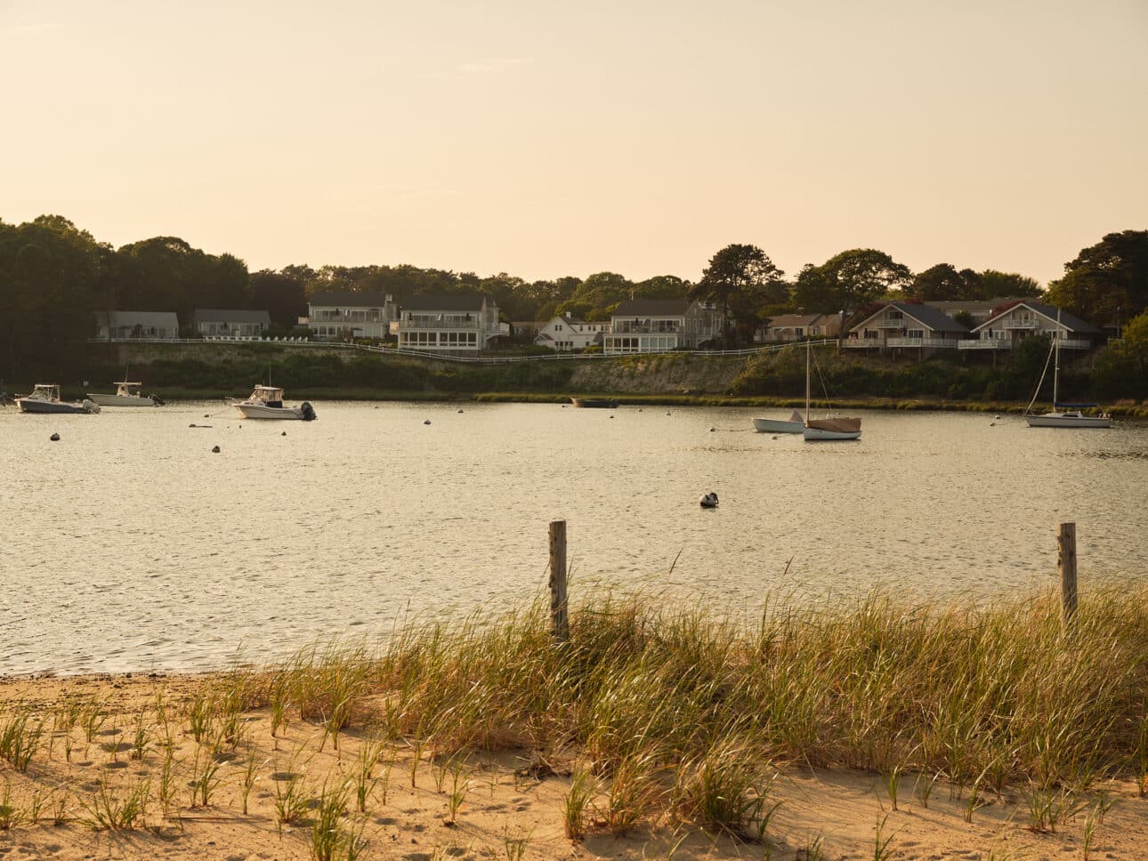 Boats are anchored in a calm bay near a sandy shore with grassy dunes. Houses line the opposite side of the water under a clear sky, evoking a tranquil scene reminiscent of the peaceful moments described in Maureen Hancock’s Soul Shift.