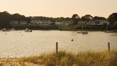 Boats are anchored in a calm bay near a sandy shore with grassy dunes. Houses line the opposite side of the water under a clear sky, evoking a tranquil scene reminiscent of the peaceful moments described in Maureen Hancock’s Soul Shift.