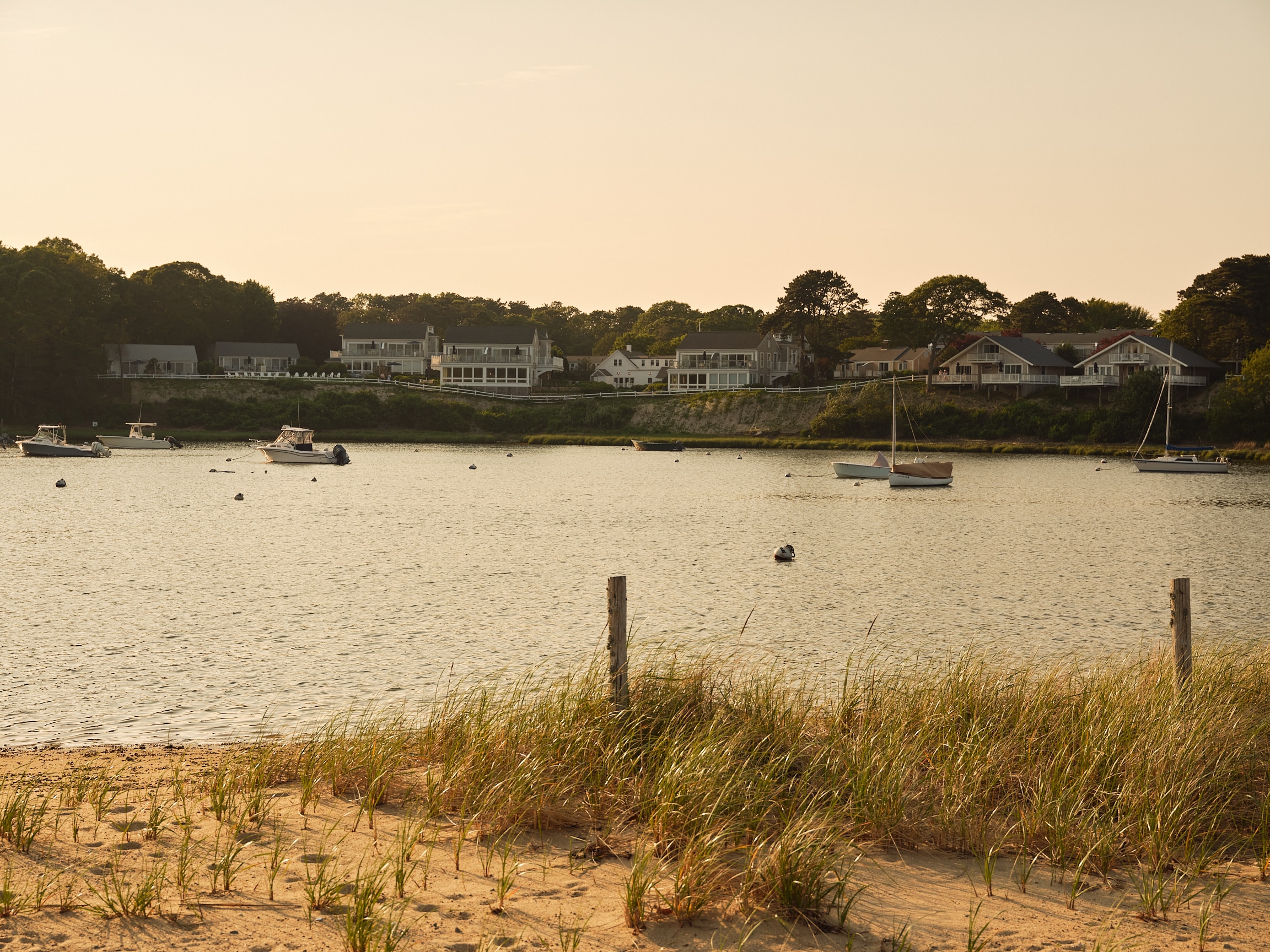 Boats are anchored in a calm bay near a sandy shore with grassy dunes. Houses line the opposite side of the water under a clear sky, evoking a tranquil scene reminiscent of the peaceful moments described in Maureen Hancock’s Soul Shift.