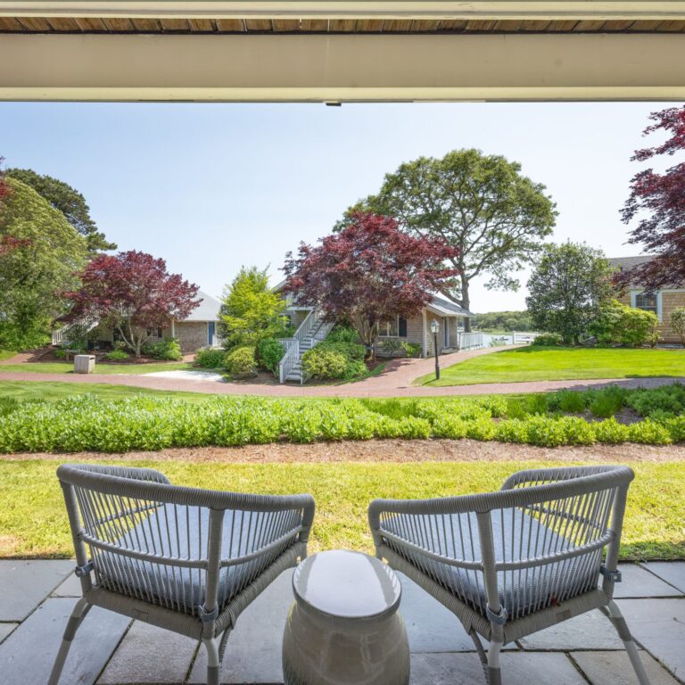 Two chairs and a small table on a patio overlook a landscaped yard with trees, shrubs, and a house in the background—perfect for relaxing after exploring a Cape Cod golf course on a sunny day.