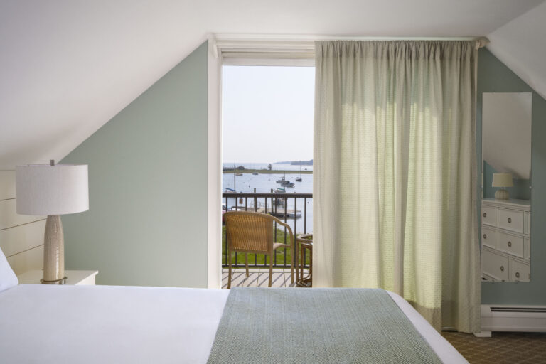A neatly made bed in a light green and white room at a Cape Cod hotel, with a balcony overlooking a waterfront dotted with boats; sheer curtains partially cover the glass door.