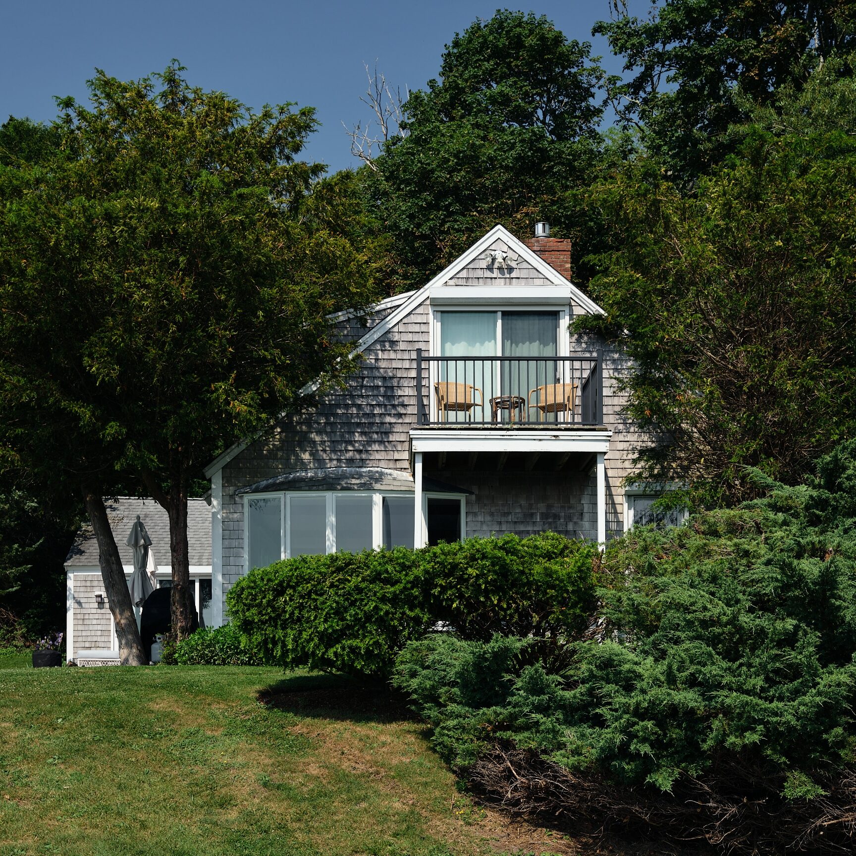 Two-story house with a shingled exterior, upper balcony with chairs, surrounded by green trees and shrubs—reminiscent of a classic Cape Cod hotel—basking in the sunlight on a beautiful day.