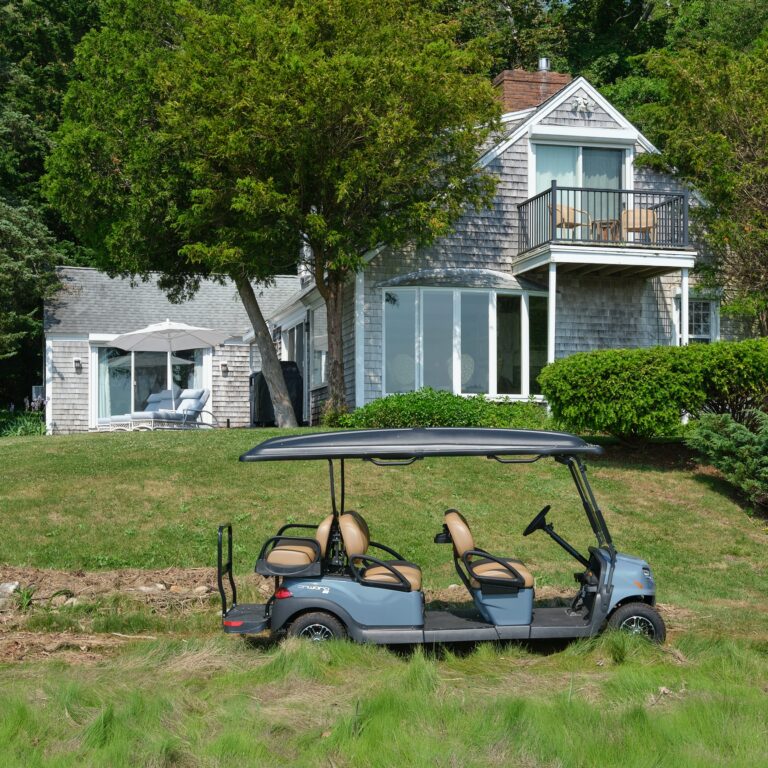 A four-seat golf cart is parked on grass in front of a grey shingled house with large windows and a balcony, surrounded by trees and shrubs at this charming Cape Cod resort.