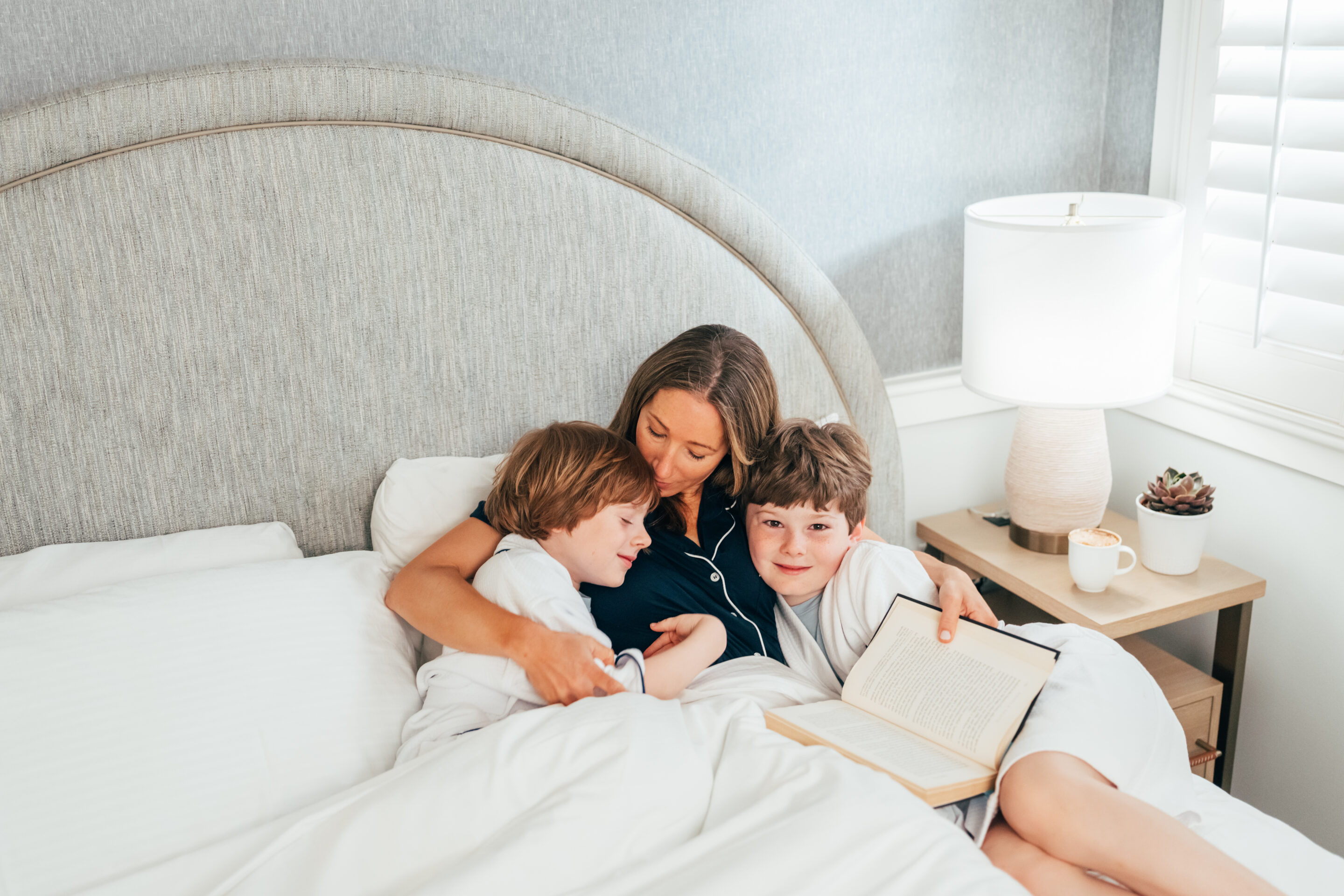 A woman sits in bed at Wequassett Resort, hugging two children; one holds an open book. A lamp, cup, and plant rest on the bedside table.