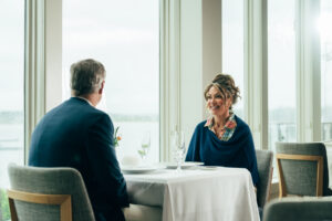 A man and a woman sit across from each other at a table for two in a bright restaurant with large windows at Wequassett Resort and Golf Club, enjoying the scenic views that Cape Cod resorts are known for.