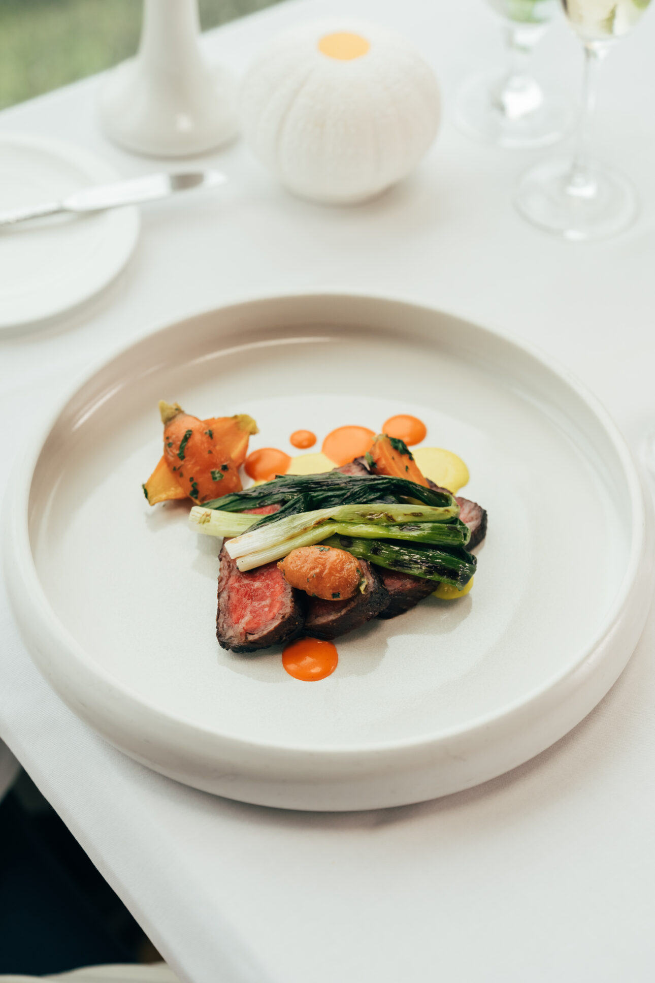 A plated dish featuring sliced steak, grilled green vegetables, roasted potatoes, and sauce dots is artfully presented at Wequassett Resort and Golf Club on a white round plate set on a white tablecloth.