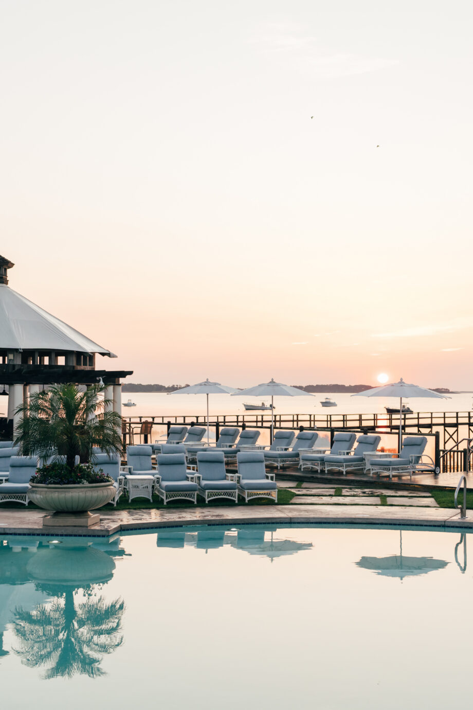 A poolside area at a Cape Cod hotel with empty lounge chairs, umbrellas, palm plants, and a gazebo, overlooking the water at sunrise or sunset.