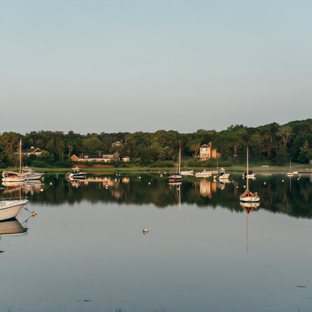 Several small boats and sailboats are anchored in a calm bay near tree-lined shores, with houses and the renowned Wequassett Resort and Golf Club visible in the background under a clear sky.