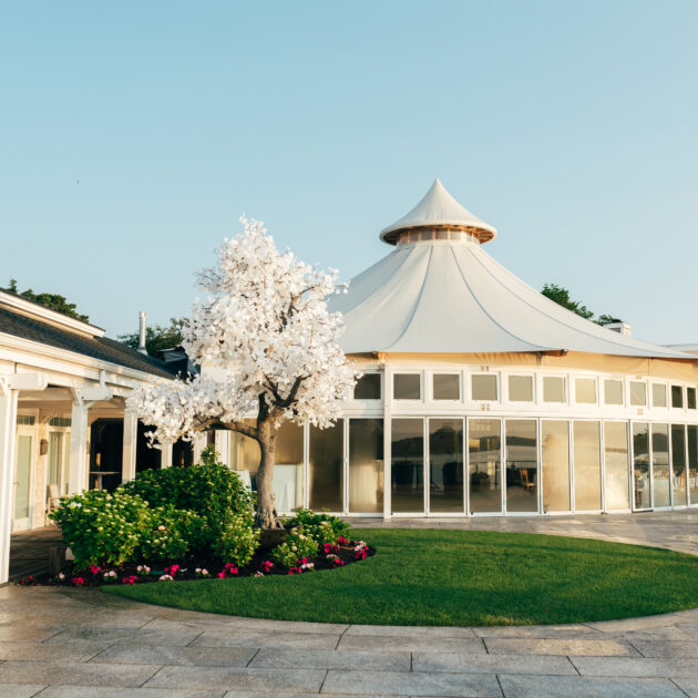 A round, glass-walled pavilion with a pointed roof sits beside a white pergola and an artificial white tree in a landscaped courtyard at Wequassett Resort.