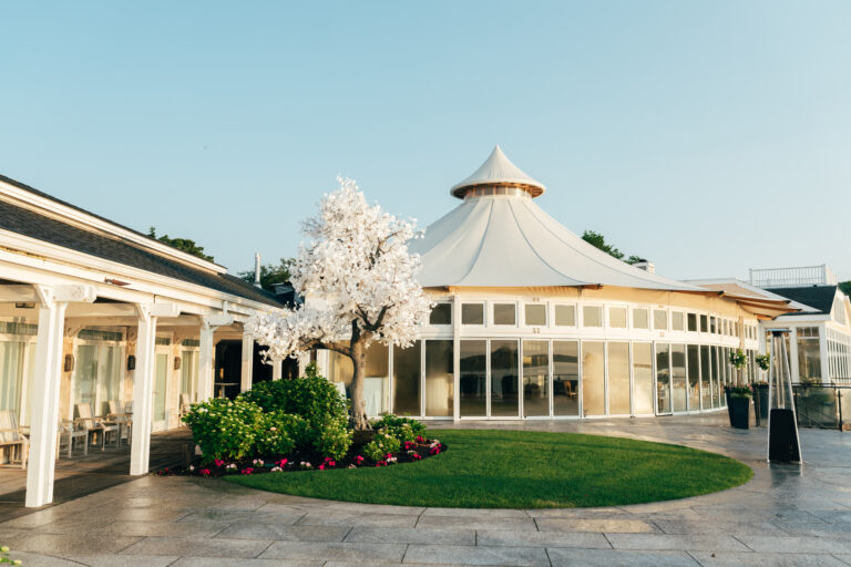 A round, glass-walled pavilion with a pointed roof sits beside a white pergola and an artificial white tree in a landscaped courtyard at Wequassett Resort.