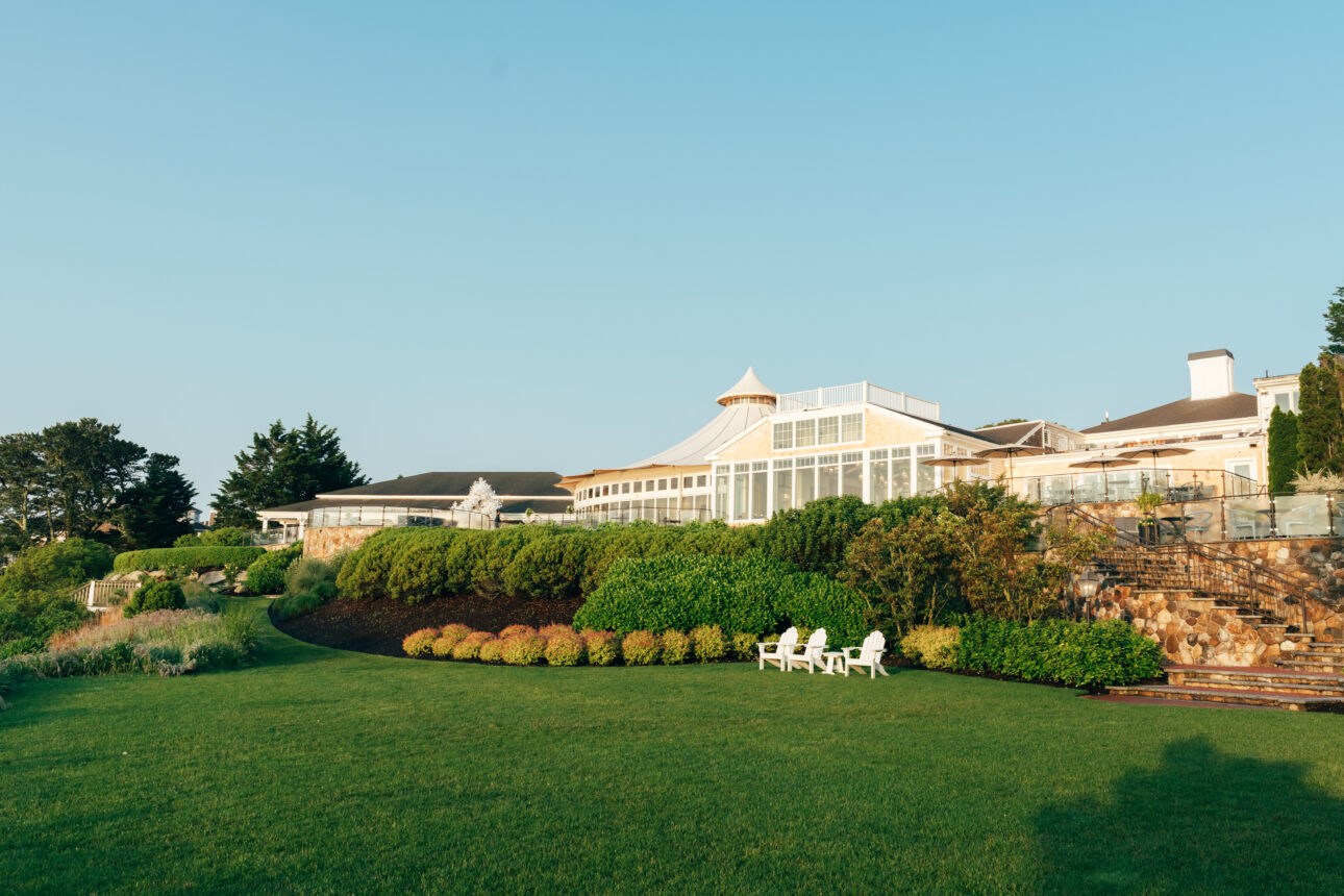 A large building with a glass-enclosed section sits on landscaped grounds with green lawns, bushes, and several white chairs—an inviting setting for events or meetings under a clear blue sky.