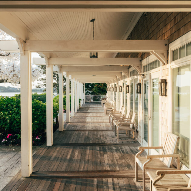 Wooden porch with white chairs lined up along glass doors at Wequassett Resort, overlooking a garden with flowers and trees, and a body of water visible in the background.