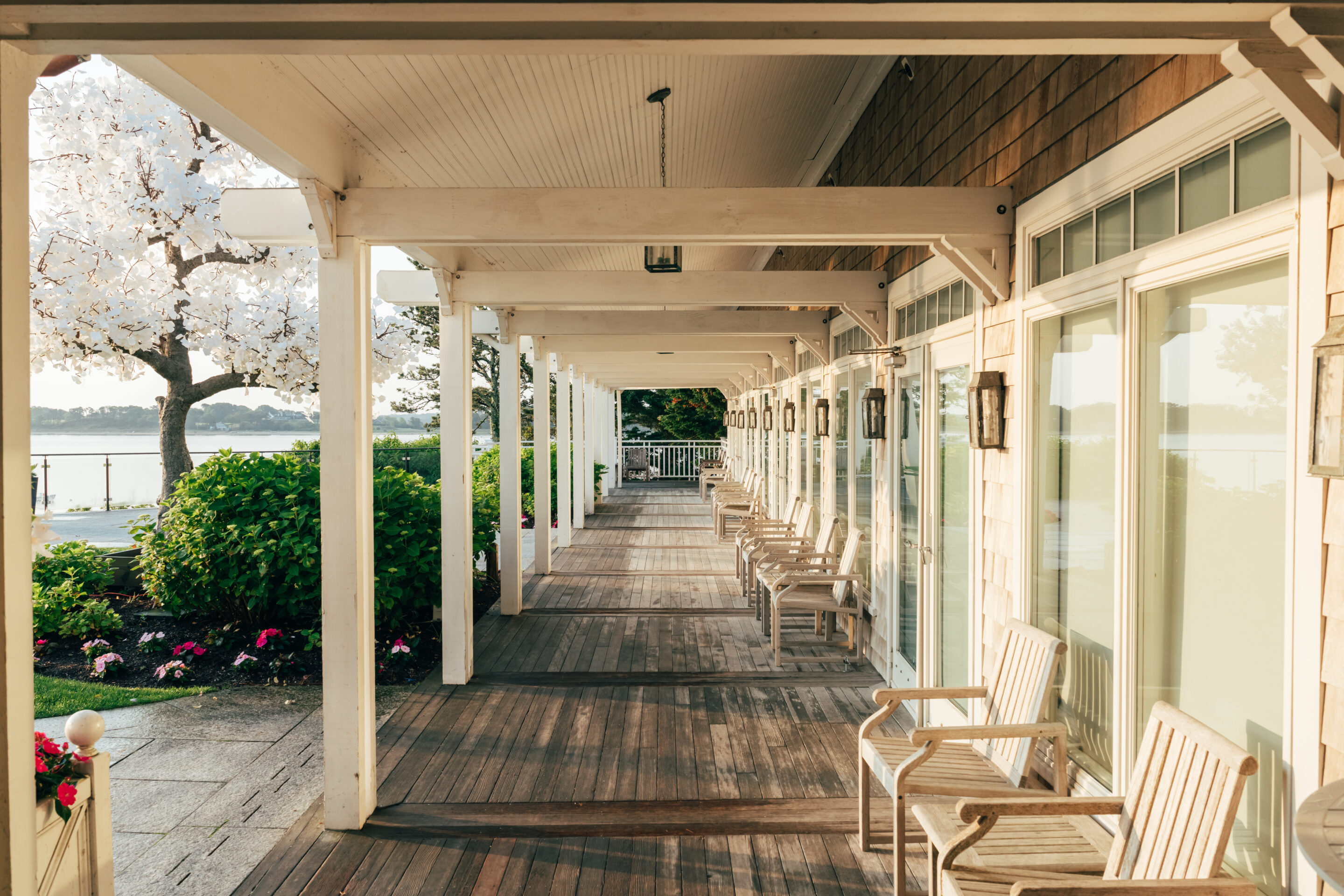 Wooden porch with white chairs lined up along glass doors at Wequassett Resort, overlooking a garden with flowers and trees, and a body of water visible in the background.