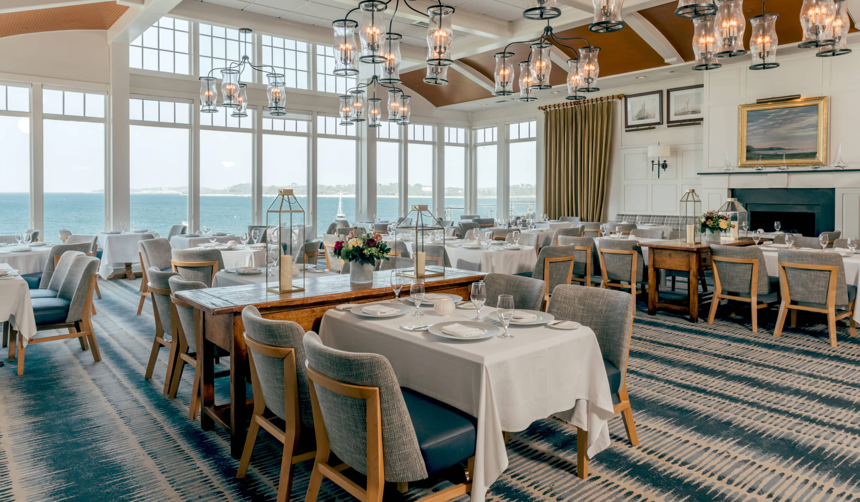 A spacious, elegant restaurant at Wequassett Resort with large windows overlooking the sea, set with white tablecloths, wooden chairs, and decorative lighting fixtures.