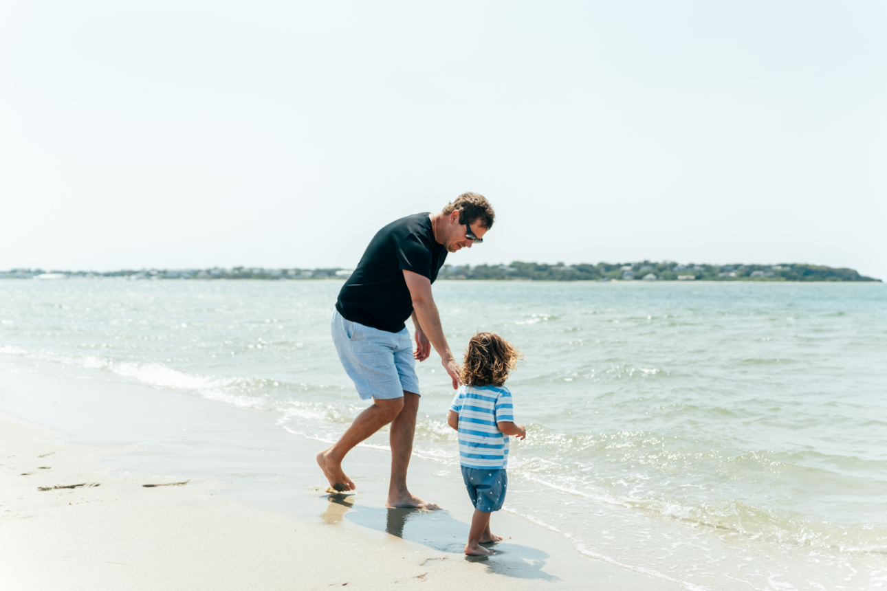 An adult and a child walk along the shoreline at a beach on a sunny day, with gentle waves and distant land in the background, capturing the serene charm found at Cape Cod resorts.